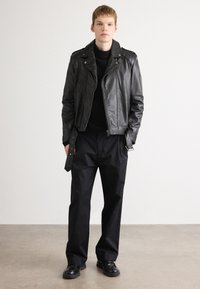 Young man standing wearing a black leather jacket, black sweater, loose black pants, and black shoes against a plain light background.