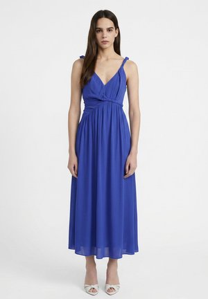 Donna con capelli lunghi e scuri che indossa un vestito midi blu reale senza maniche con spalline da annodare e tacchi bianchi aperti, in piedi davanti a uno sfondo semplice.