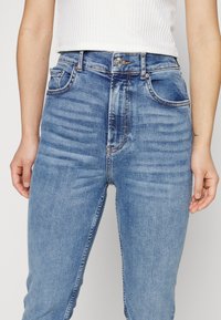 Blå denim jeans med hög midja, fem fickor och subtila blekningar. Kontraststygn och en detalj med knappknäppning är synliga.