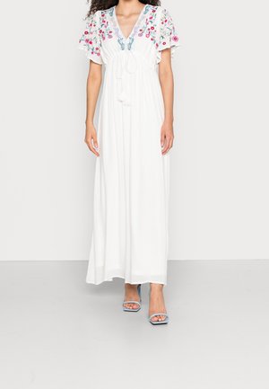 Robe maxi blanche à manches volantes, broderie florale sur les épaules, col en V avec liens à pompons, portée avec des talons ouverts bleu clair.