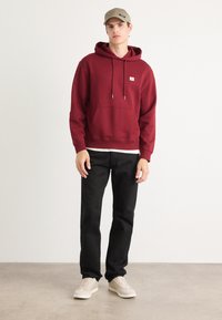 Felpa con cappuccio color marrone bordeaux, con tasca frontale, cordini bianchi, pantaloni neri e sneakers beige. Tessuto liscio, vestibilità casual.