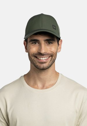 Lächelnder Mann mit kurzen dunklen Haaren, der eine olivgrüne Baseballkappe und ein cremefarbenes T-Shirt mit Rundhalsausschnitt vor hellem Hintergrund trägt.