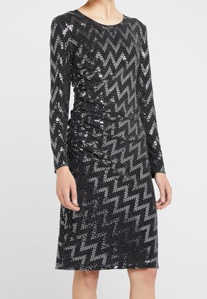 Schwarzes knielanges Kleid mit langen Ärmeln, silbernen Zickzack-Pailletten und einem gerafften Taillendetail an einer stehenden Person.