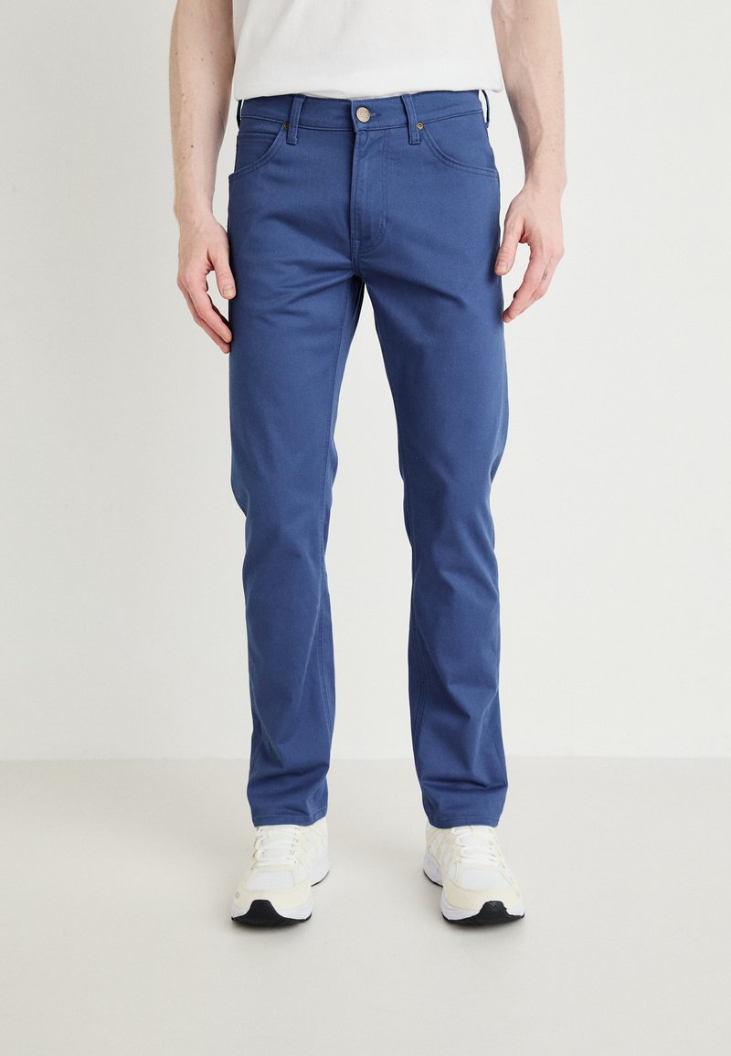 Modré bavlnené džínsy so Slim fit strihom, obsahujúce päť vreciek a zips s gombíkovým zapínaním. Nosené s bielymi atletickými topánkami.