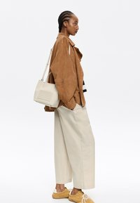 Giacca in suede marrone, pantaloni beige a gamba larga, scarpe gialle e una borsa color crema con tracolla. Design semplice e funzionale.