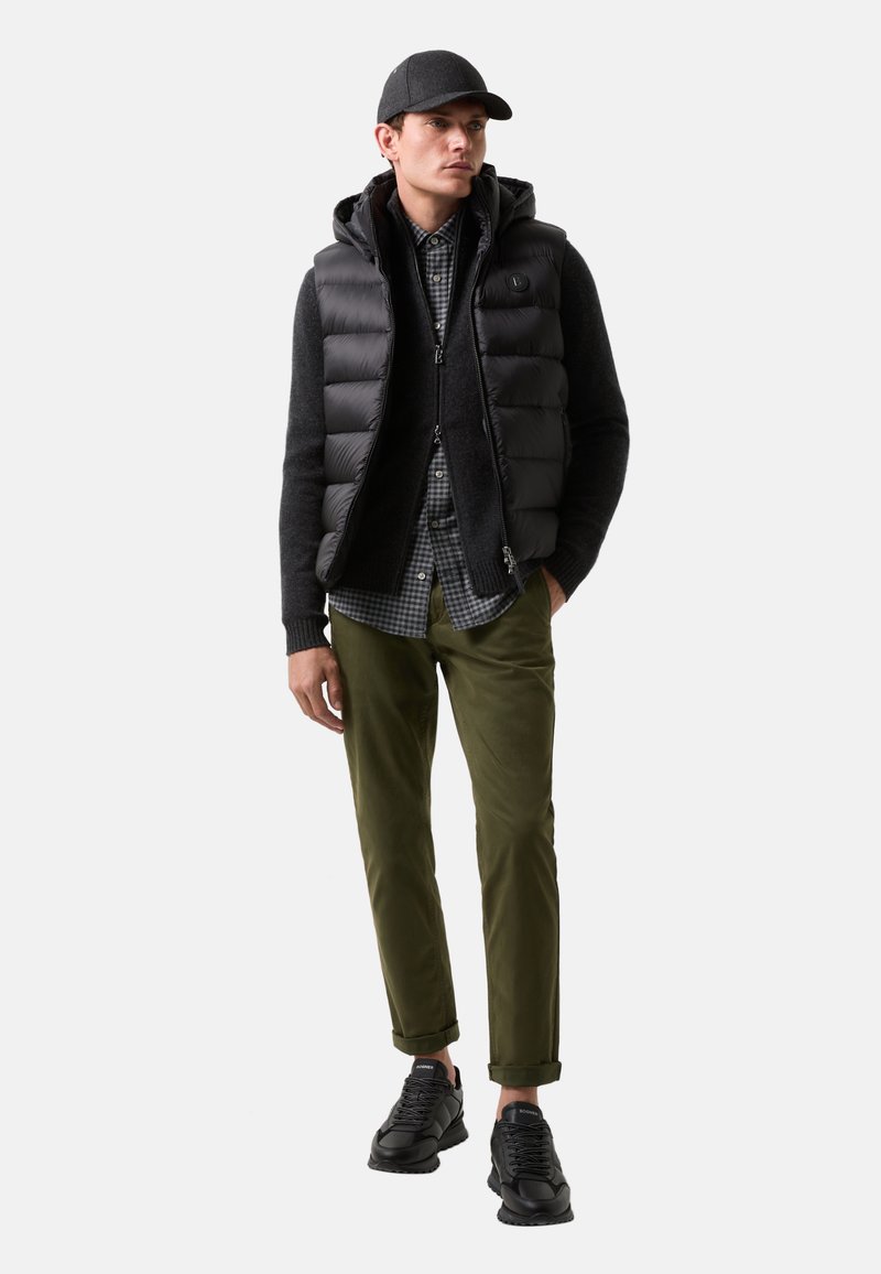 Schwarze Steppweste über einem dunklen Pullover, kombiniert mit einem karierten Hemd, olivgrünen Hosen und schwarzen Sneakers. Einschließlich einer grauen Kappe.