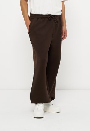 Egy férfi sötétbarna bőszárú, megkötős nadrágot és fehér tornacipőt visel, egyik kezét zsebében tartva áll fehér háttér előtt.