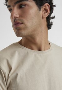 Beige színű pamut póló kerek nyakkivágással és sima textúrával, oldalról látható modellen. Finom varrások látszanak.