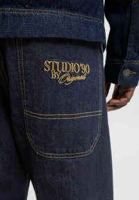 Chaqueta de mezclilla oscura con costuras amarillas y un logo bordado "STUDIO'90 BY Originals" en el bolsillo trasero. Detalle de botones visible.