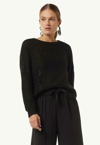 Schwarzer Strickpullover mit lässiger Passform, verziert mit subtilen Pailletten, Rundhalsausschnitt und langen Ärmeln. Kombiniert mit weit geschnittenen schwarzen Hosen.