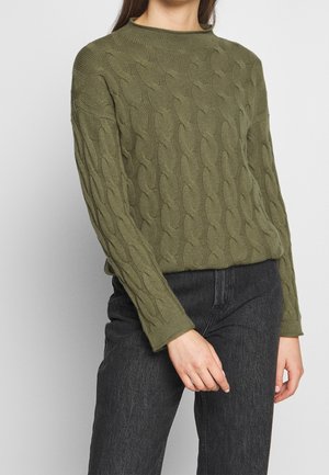 Femme portant un pull en maille torsadée vert olive à manches longues et un jean noir taille haute, debout devant un fond uni.