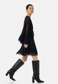 Robe noire à plusieurs couches avec des manches semi-transparentes, présentant un ourlet évasé. Associée à des bottes en cuir noir montantes et à une texture lisse.