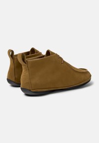 Rudos zomšos casual slip-on batai su juodais padais, nugaros traukimo kilputėmis ir minimalistiškais raišteliais, ant neutraus fono.