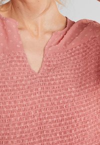 Blouse rose texturée avec un design smocké, décolleté en V et motif à pois sur la partie supérieure. Tissu léger au toucher doux.