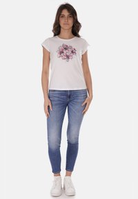 Maglietta bianca a maniche corte con design floreale, abbinata a jeans aderenti blu chiaro e sneakers bianche. Outfit casual.