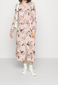 Žena v růžových květinových šatech midi s knoflíkovým zapínáním a páskem v pase, bíl�ých teniskách, ponožkách a malou bílou kabelkou přes rameno.