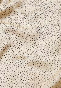 Tissu crème avec de petites taches noires irrégulières dispersées, montrant des plis doux et une texture.