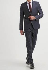 Granatowy garnitur na miarę z dopasowaną marynarką i spodniami z prostym przodem. Uzupełniony białą koszulą i bordowym krawatem w kropki, wykończony czarnymi butami.