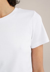 Balta medvilnė marškinėliai su apvaliu kaklu ir trumpomis rankovėmis, pasižymintys lygaus audinio savybėmis ir paprastu dizainu be grafinių elementų.