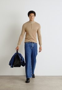 Maglione beige con zip, jeans ampi blu, stivali neri, che porta una giacca blu. Texture liscia, design casual, dettaglio di logo minimo sul maglione.