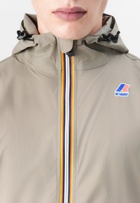Chaqueta impermeable de color crema claro con capucha, que presenta rayas verticales multicolores y un logo de K-Way en el pecho. Textura de tela suave.