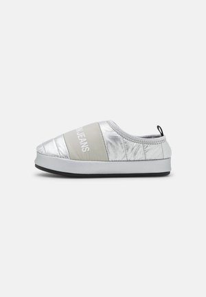 Harmaalla "JEANS" -tekstillä varustettu hopeanvärinen, pehmustetulla tikatulla päällisellä varustettu slip-on-tennari, valkoisella pohjalla ja mustalla kuvioinnilla yksinkertaisella taustalla.
