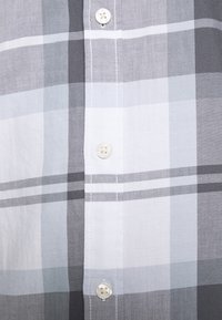 Primo piano di un tessuto a quadri grigio, bianco e azzurro chiaro con bottoni bianchi cuciti verticalmente sul davanti di una camicia.