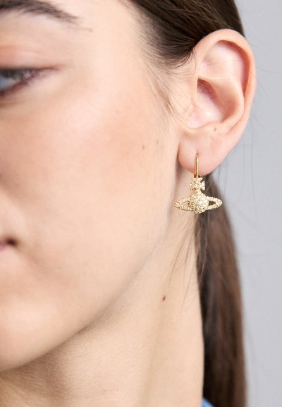 Zlaté kruhové náušnice se spirálovým designem, zdobené malými, čirými krystaly, zobrazené na uchu modelky. Hladký povrch, lesklý finiš.