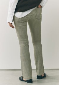 Karierte, ausgestellte Hose in Grün und Beige mit glatter Textur; kombiniert mit einem weißen Hemd und schwarzen flachen Schuhen.