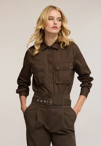 Hnědá košile s límečkem a dvěma náprsními kapsami, doplněná oknoflíky na patent. Kombinováno s hnědými kalhotami s vysokým pasem a páskem.