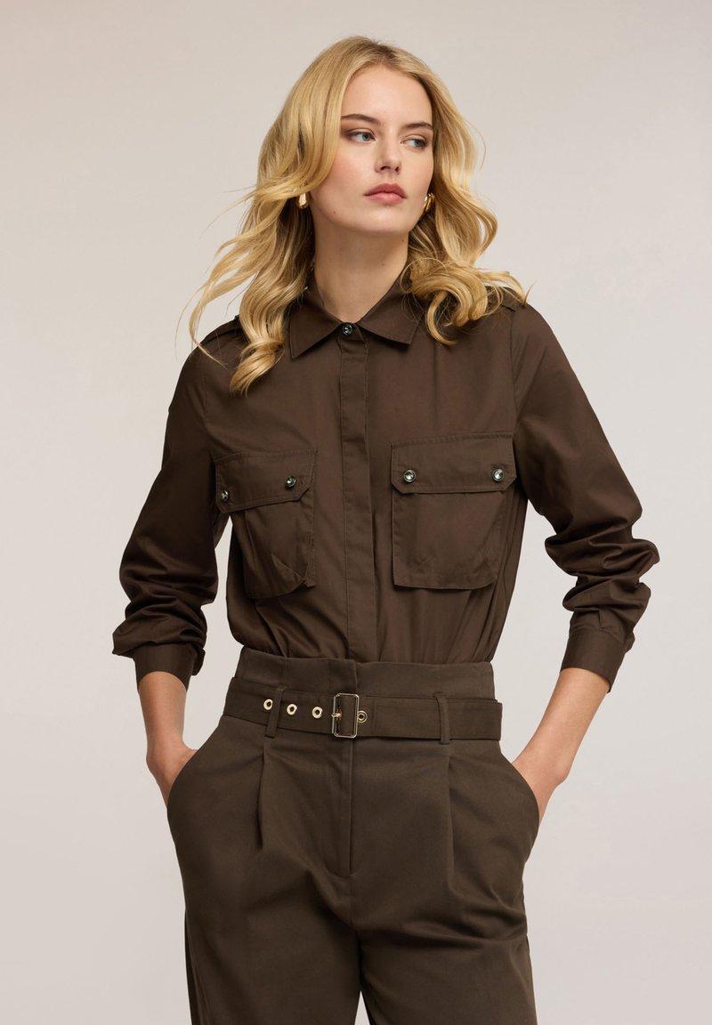 Hnědá košile s límečkem a dvěma náprsními kapsami, doplněná oknoflíky na patent. Kombinováno s hnědými kalhotami s vysokým pasem a páskem.