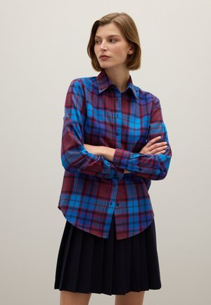 Camisa de cuadros en tonos azules, morados y rojos; de manga larga; con dobladillo curvado; combinada con una falda negra plisada.