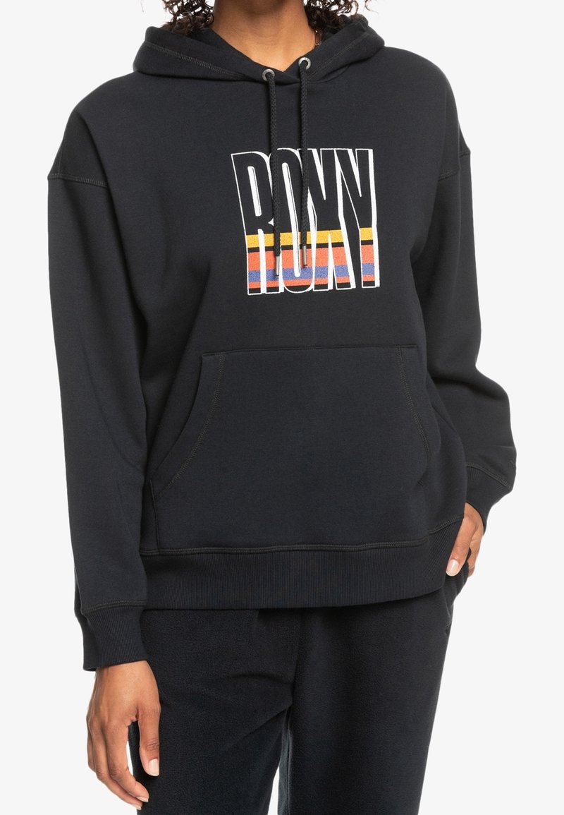 Personne portant un sweat à capuche noir avec une poche frontale et le texte « ROXY » en blanc avec des bandes multicolores sur la poitrine.