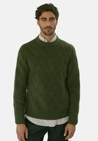 Pull en tricot vert olive avec un motif de losanges texturé, col rond et poignets côtelés, superposé à une chemise blanche à rayures.