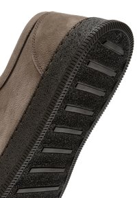 Chaussure en suede marron avec une semelle en caoutchouc noir texturé, comprenant des rainures et des crêtes horizontales pour une meilleure adhérence. Finition cousue sur le côté.