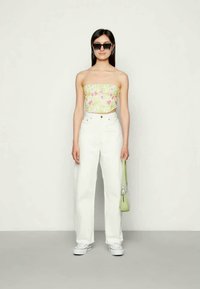 Mulher a usar óculos de sol, top tubular floral, calças brancas de cintura alta, sapatilhas brancas, a segurar uma pequena bolsa verde, de pé contra um fundo liso.