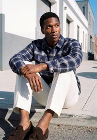 Homme portant une chemise à carreaux bleue, un pantalon blanc et des mocassins marron assis sur un rebord de trottoir, regardant sur le côté à la lumière du jour.