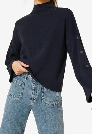 Kobieta ubrana w granatowy prążkowany sweter z guzikami na rękawach oraz jasnoniebieskie, wysokie jeansy z naszywanymi przednimi kieszeniami.