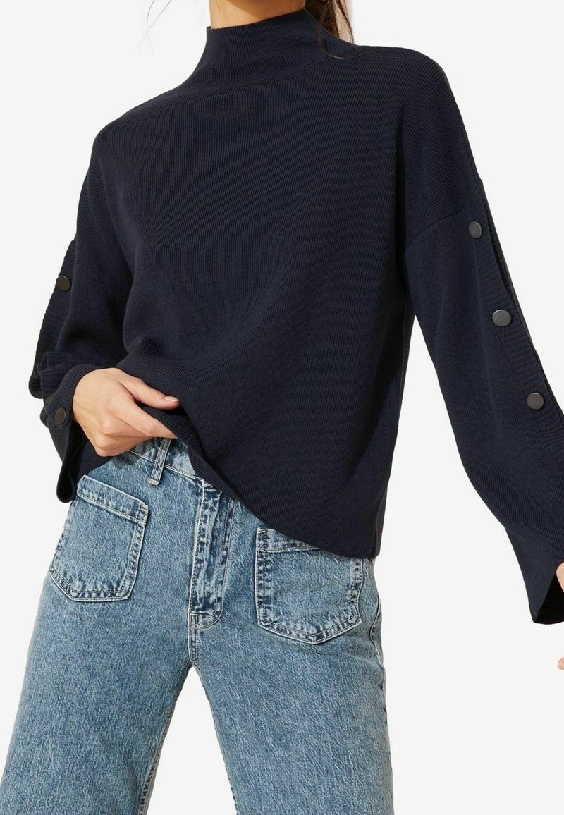 Femme portant un pull côtelé bleu marine avec des boutons sur les manches et un jean taille haute bleu clair avec des poches plaquées devant.
