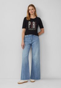 T-shirt noir à manches courtes avec "PUR DELICE" imprimé en lettres colorées, associé à un jean large bleu clair et des baskets blanches.