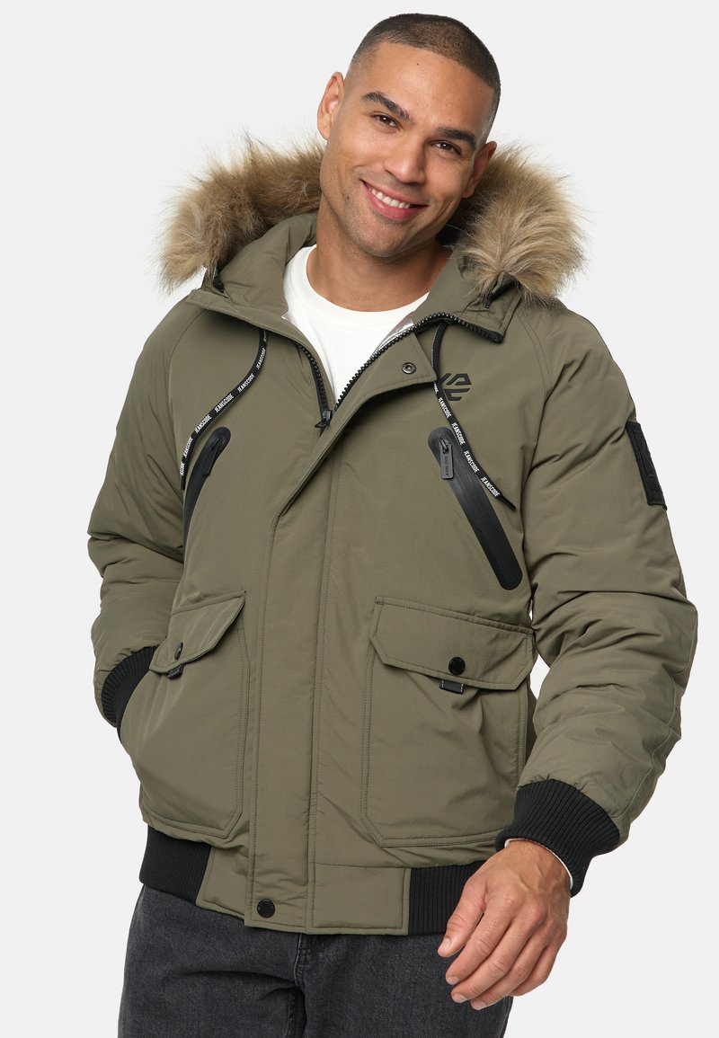 Olivgrüner Parka mit einem mit Fell gefütterten Kapuze, mehreren Reißverschlusstaschen, Rippbündchen und einem schwarzen Logo-Detail auf der Brust.