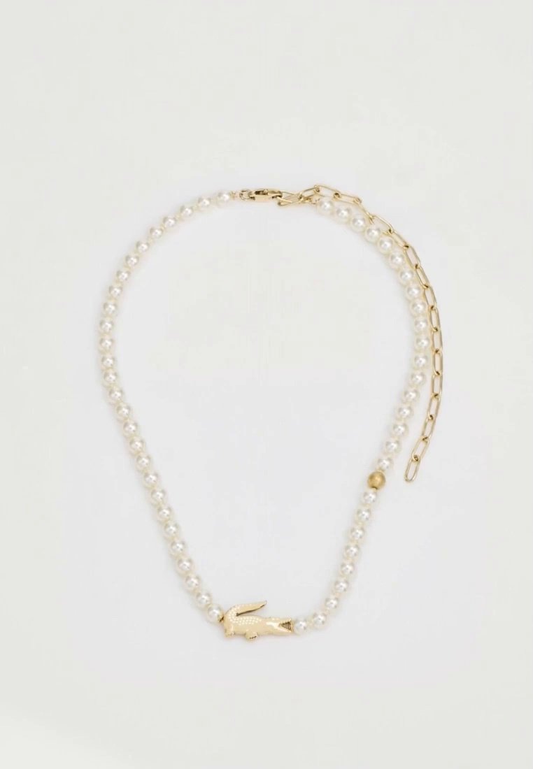 Un collier de perles blanches doté d’un fermoir doré en forme de crocodile et d’une chaîne dorée réglable repose sur un fond clair.