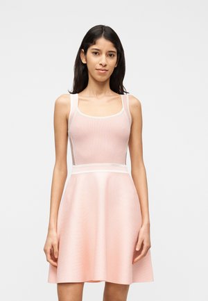 Młoda kobieta o długich ciemnych włosach, ubrana w różową, dopasowaną sukienkę bez rękawów z rozkloszowanym dołem, stojąca na tle jednolitego białego tła.