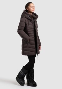 Brun puffy jakke med høj krave, lynlåse og snoretræk i hætten. Sammen med sorte leggings og isolerede sorte sneskøjtter.