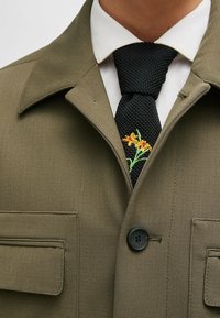 Olijfgroene jas met een gestructureerde afwerking, grote revers en twee borstzakken. Zwarte stropdas met geborduurde gele bloemen en groene stengels.