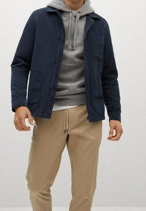 Uomo che indossa una felpa grigia con cappuccio sotto una giacca blu scuro con bottoni e pantaloni beige con coulisse, in piedi davanti a uno sfondo neutro.