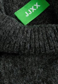 Sweater de malha cinza escuro com uma superfície texturizada, apresentando uma etiqueta verde proeminente rotulada "JJXX". A malha tem detalhes canelados.