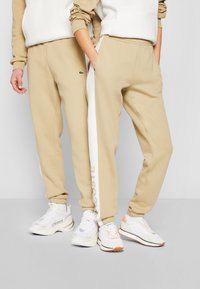 Beige sweatpants med elastiska muddar, med en vit sidostrand och Lacoste-logotyp. Matchas med vita träningssneakers.