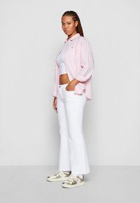 Rosa, knäppt skjorta över en vit crop top; vita utsvängda jeans; guld- och vita sneakers; flätad frisyr; stående pose.