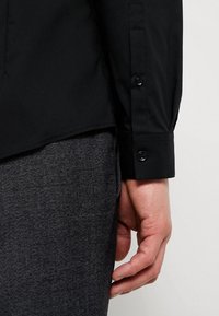 Zwarte shirt met een gladde textuur, met een knoopmanchet, gecombineerd met grijze textuur broek. Focus op de detail van de mouw en hand.